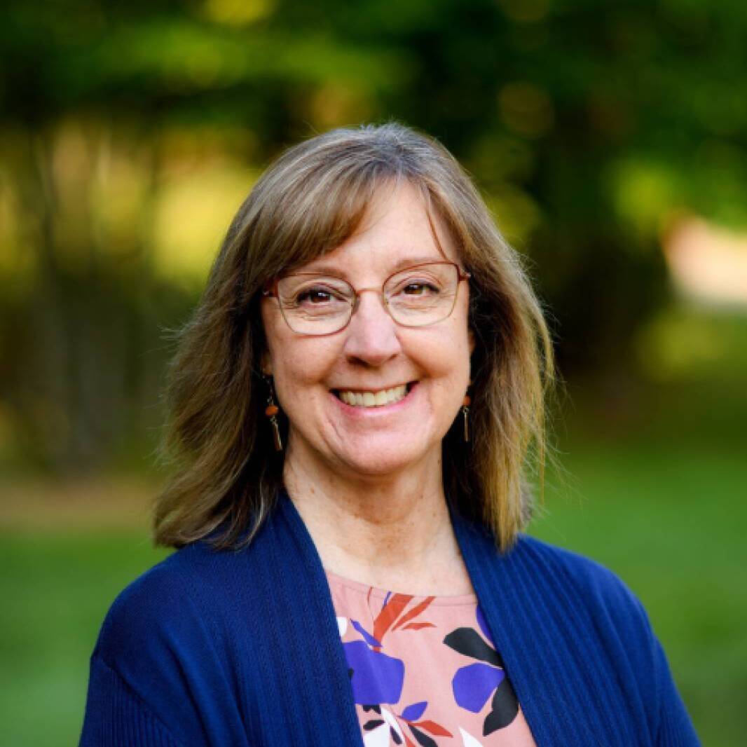 A woman with bangs and short brown hair, glasses, a blue blazer, and floral pink, blue, orange, and black blouse smiles at the camera in front of an outdoor green grass background